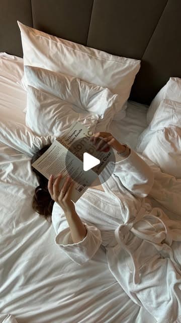 A woman in a white robe reading a magazine in bed with white linens